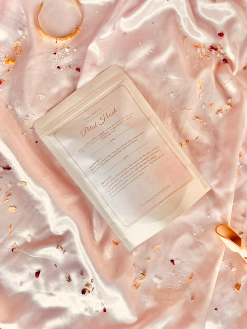 A stand-up pouch package labeled 'Petal Hush' lies on shiny pink satin fabric with scattered dried flower petals and small decorative gold-toned accessories around it.
