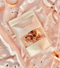 A stand-up pouch labeled 'Petal Hush' by LUMA Bath Co, placed on shiny pale pink satin fabric with scattered dried petals and small decorative accents.