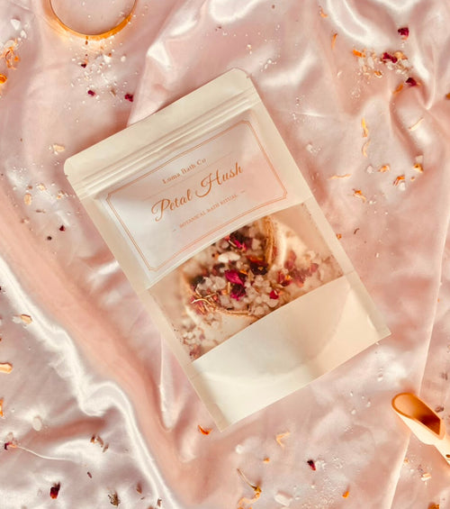 A stand-up pouch labeled 'Petal Hush' by LUMA Bath Co, placed on shiny pale pink satin fabric with scattered dried petals and small decorative accents.