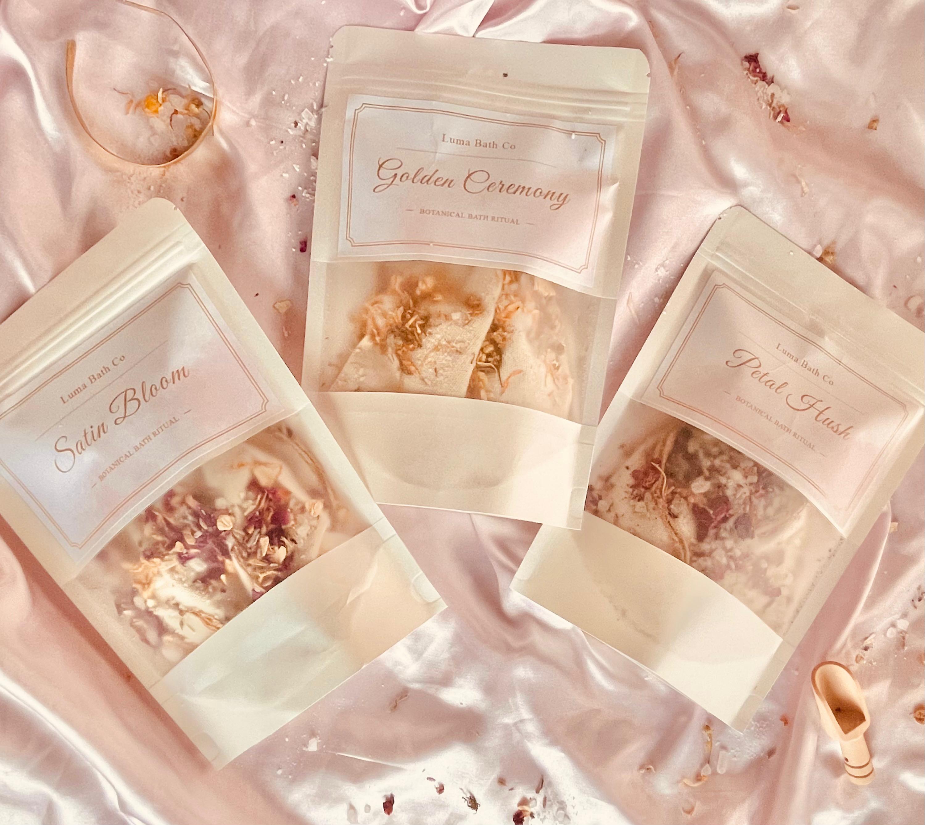 Three stand-up pouches of bath salts arranged on shiny pink satin fabric. The pouches are labeled 'Luma Bath Co' and feature the names 'Satin Bloom,' 'Golden Ceremony,' and 'Petal Hush,' with clear windows showing the bath salts inside.